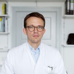 Leonhard Schilbach steht in Arztkittel vor einem weißen Bücherregal